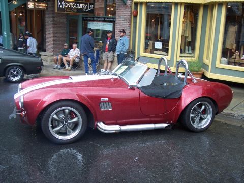 Red Cobra-style roadster with white stripes and side pipes parked on wet street