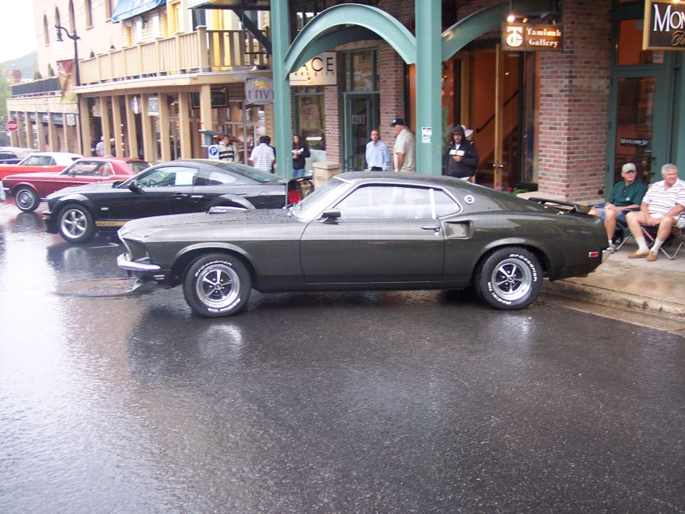 Olive Ford Mustang fastback in side view on a wet city street with onlookers