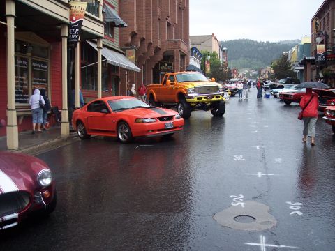 Red Mustang coupe and lifted orange pickup at a rainy street car show