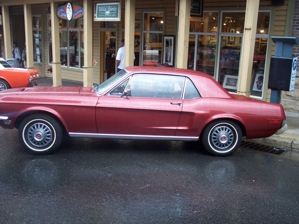 Red Ford Mustang hardtop coupe parked in profile on wet street by storefront