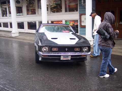 Black Ford Mustang with white hood stripe on a wet street, people walking past shops.