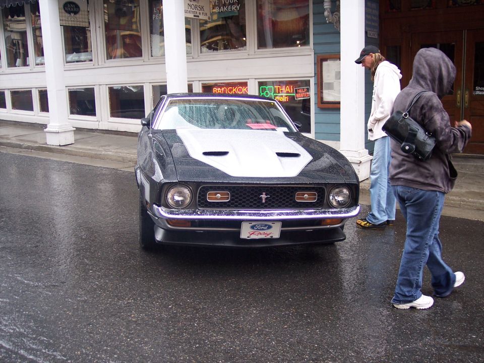 Black Ford Mustang with white hood stripe on a wet street, people walking past shops.