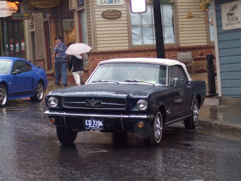 Dark first-gen Ford Mustang convertible with white top on a wet street, front 3/4 view.