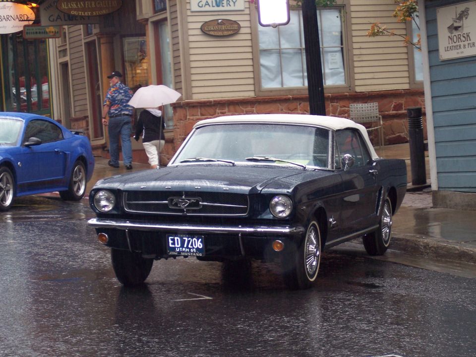 Dark first-gen Ford Mustang convertible with white top on a wet street, front 3/4 view.