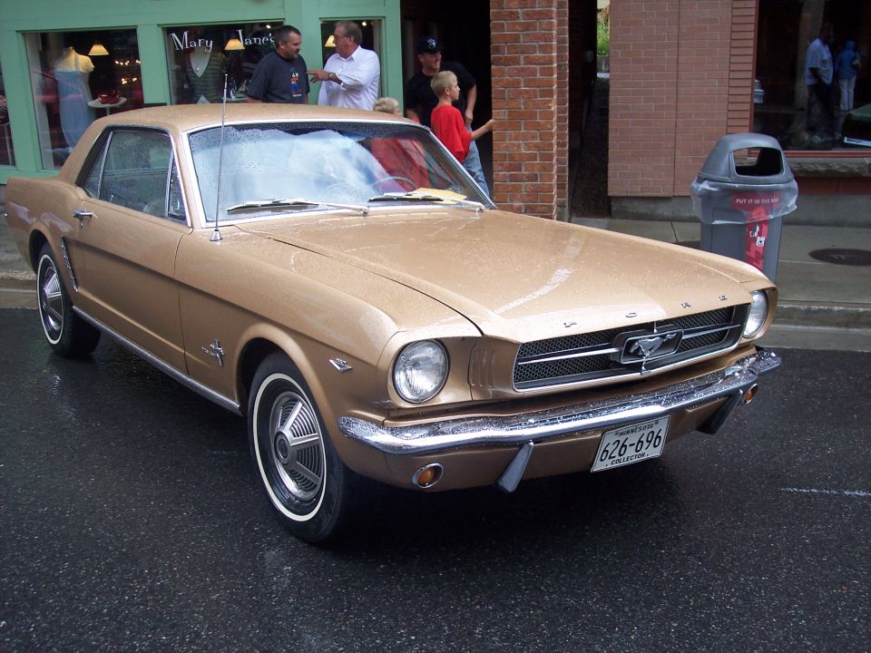 Gold 1960s Ford Mustang coupe, front 3/4 view, on wet street with chrome bumper and whitewalls.