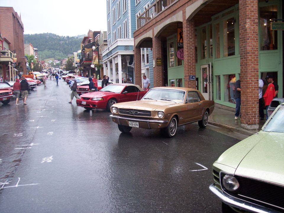 Gold classic Ford Mustang coupe parked on a wet street at a car show, with a red car behind.