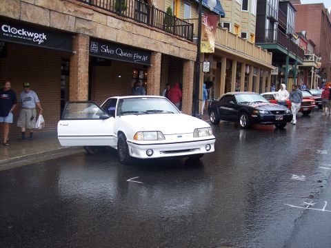 White Fox-body Ford Mustang with open door on a wet downtown street with shops and other cars
