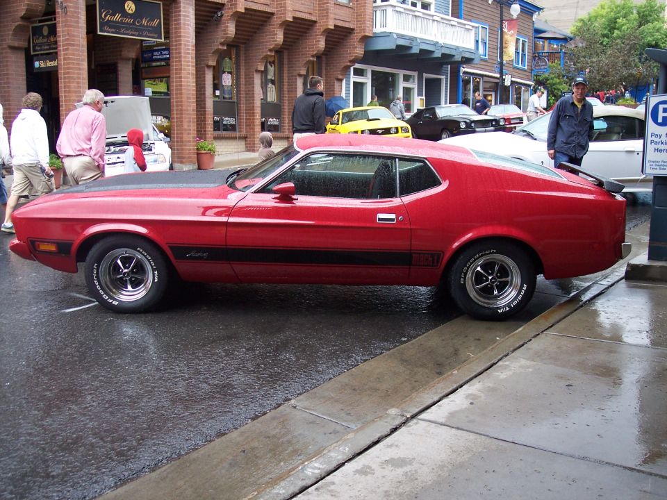 Red Mustang Mach 1 fastback in side profile on a wet street at a car show