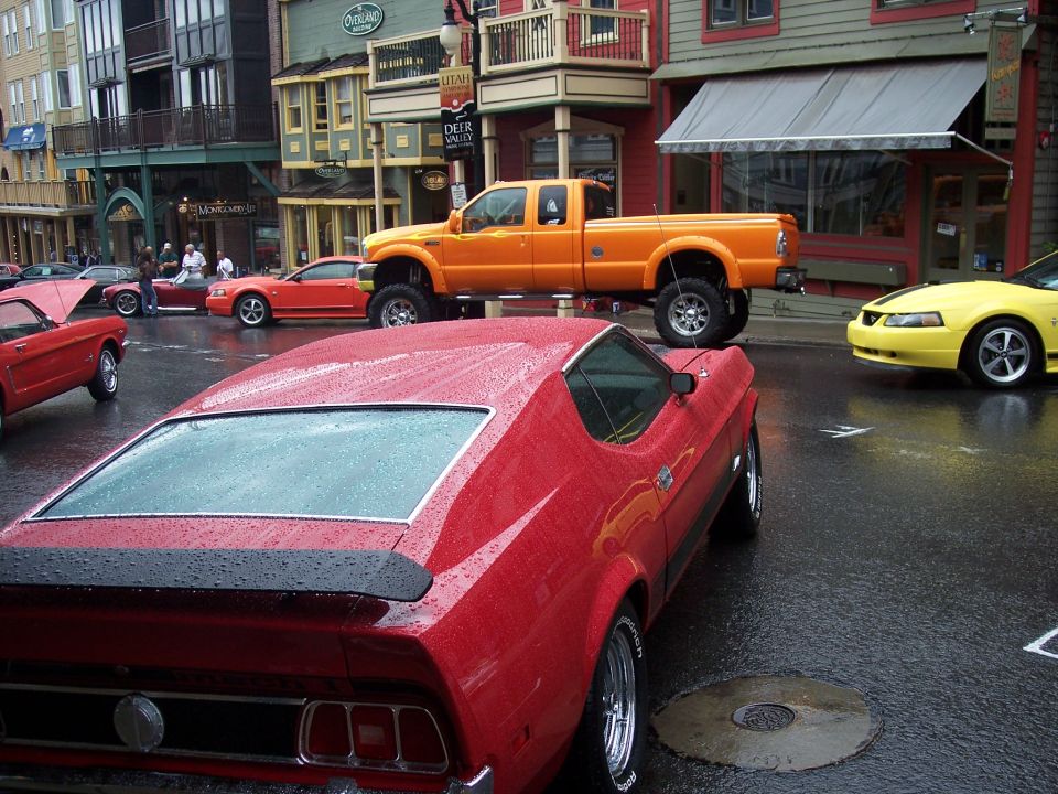 Red fastback muscle car rear 3/4 view on wet street with orange lifted truck and yellow car behind.