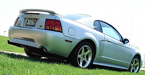 Silver Ford Mustang Cobra rear view, parked on grassy hill. COBRA lettering on trunk lid