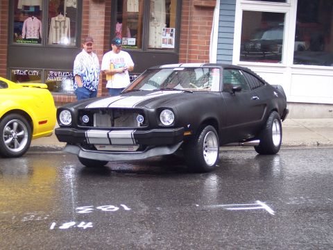 1972 Ford Ranchero - A black coupe with white racing stripes is parked on a wet street next to a yellow car.