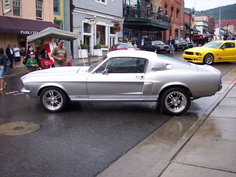 1972 Ford Ranchero - A silver 1967 Ford Mustang Fastback is parked on a wet street in front of shops.