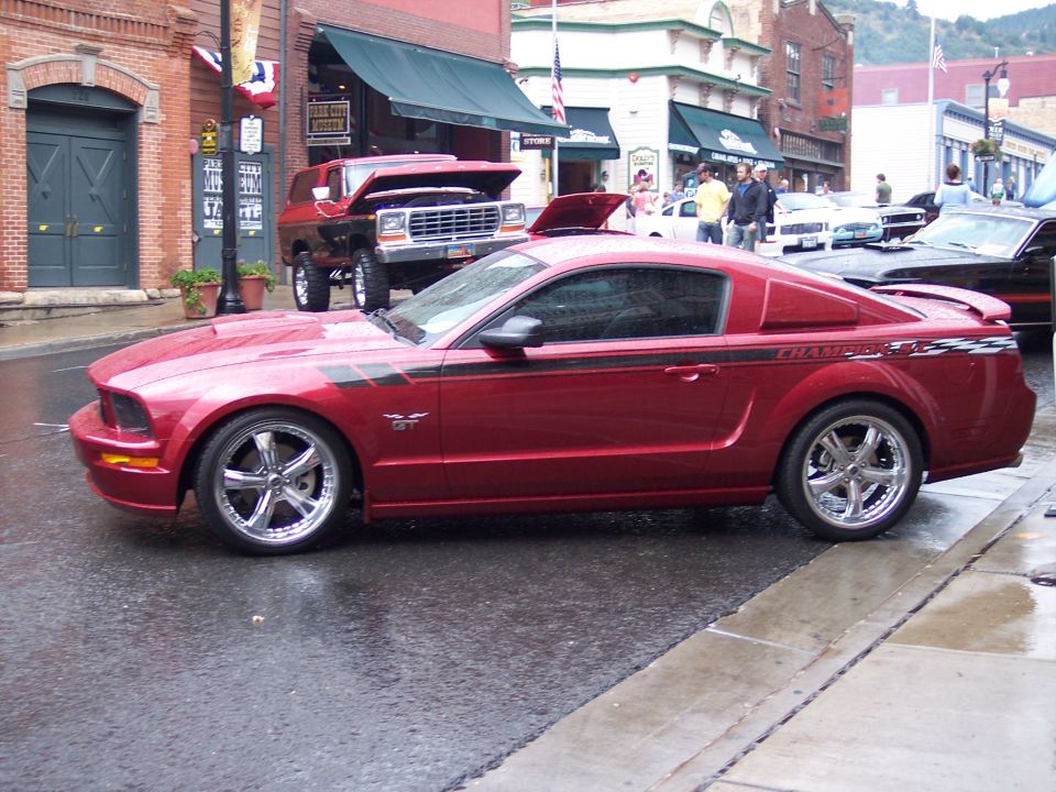 Red Ford Mustang GT coupe with black stripes, parked on wet street. Another vehicle, possibly a red SUV, is behind it