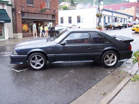 1972 Ford Ranchero - A dark gray coupe with a spoiler is parked on a wet street in front of brick buildings.
