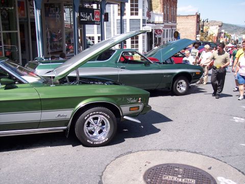 1972 Ford Ranchero - Two green Rancheros are displayed with hoods open on a street lined with shops.