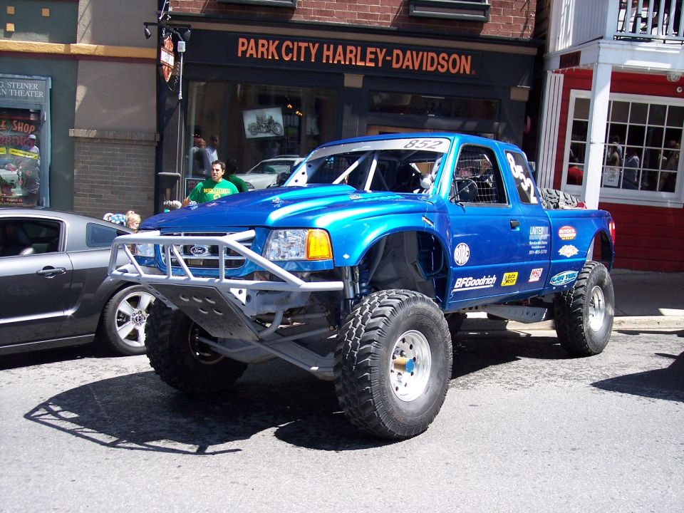 Blue off-road Ford Ranger pickup truck with large tires and roll cage, parked next to a gray car. Building in background says 'PARK CITY HARLEY-DAVIDSON'