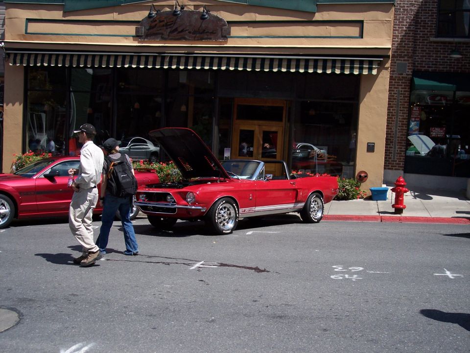 Red 1968 Ford Mustang convertible parked on street, hood open. Another red car is parked to the left