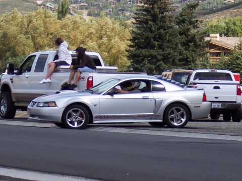 1972 Ford Ranchero - A silver coupe with racing stripes is parked next to two pickup trucks, with people sitting in the beds of the trucks.