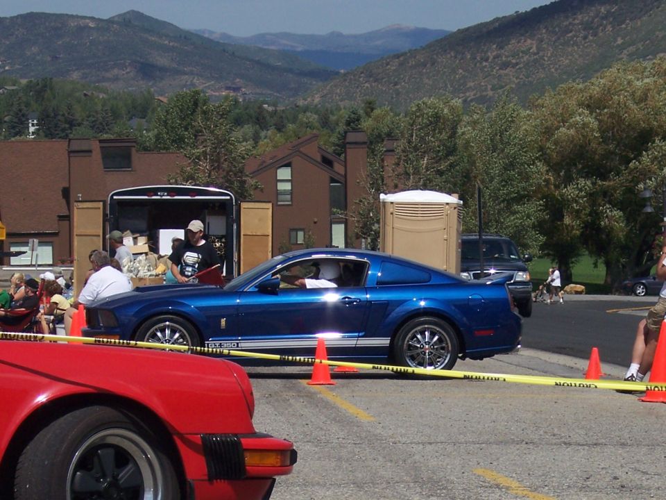 Blue Ford Mustang parked next to a trailer, with mountains in the background. A red car is in the foreground
