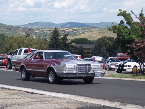 1972 Ford Ranchero - A 1977 Ford Ranchero pickup truck is seen driving, with other vehicles parked in a lot behind it.