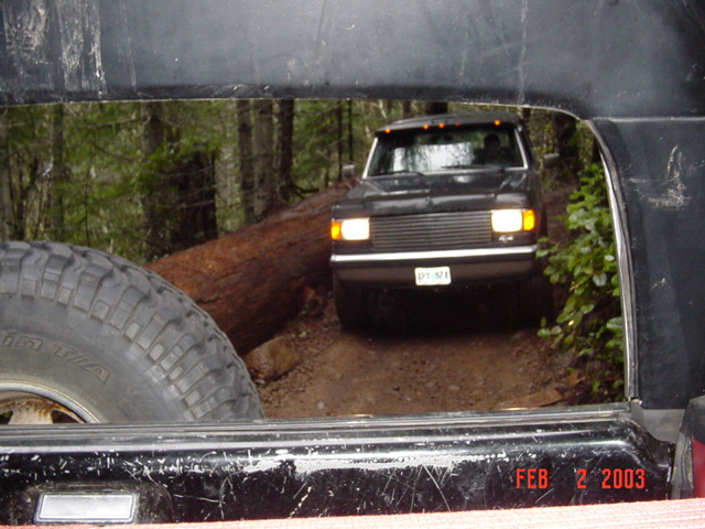 View through a vehicle's rear window of a dark Chevy truck crossing a log on a forest trail