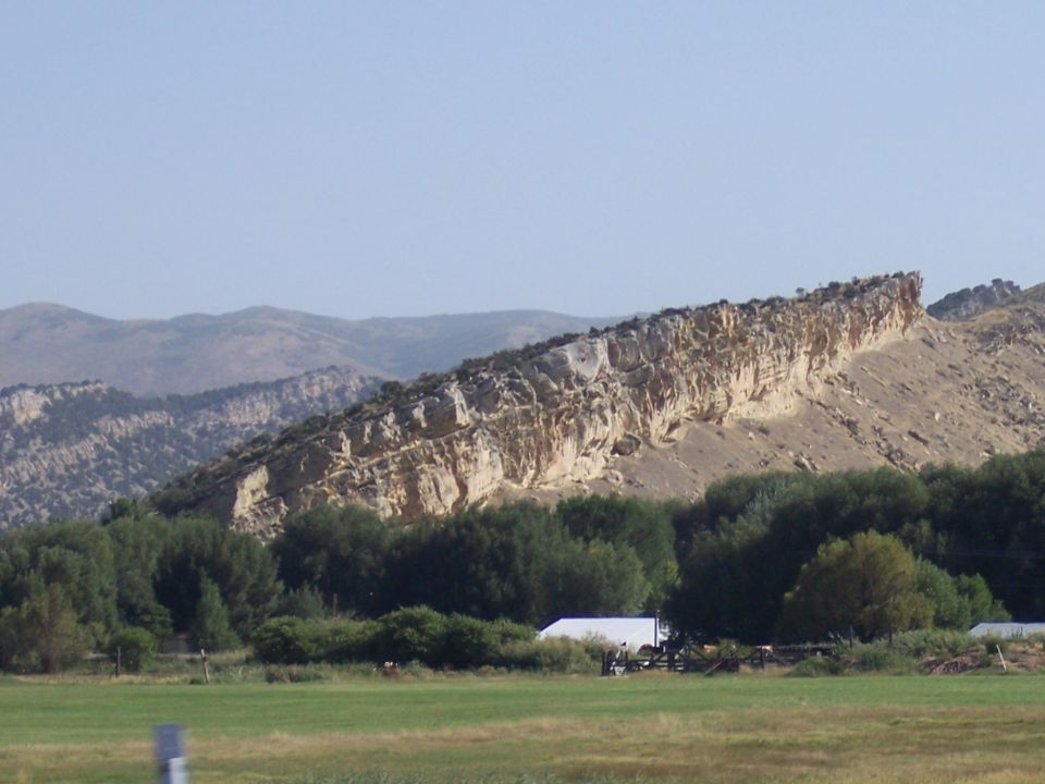 Long, layered rock formation overlooks trees and a distant white building