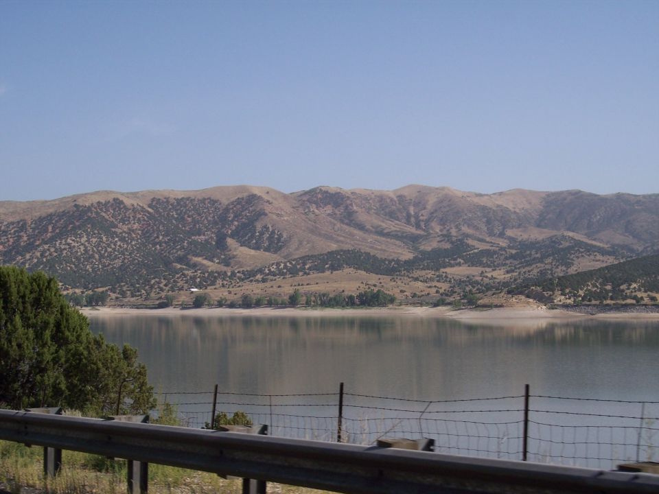 Mountain range reflected in a calm lake under a clear blue sky, with trees and a fence in the foreground