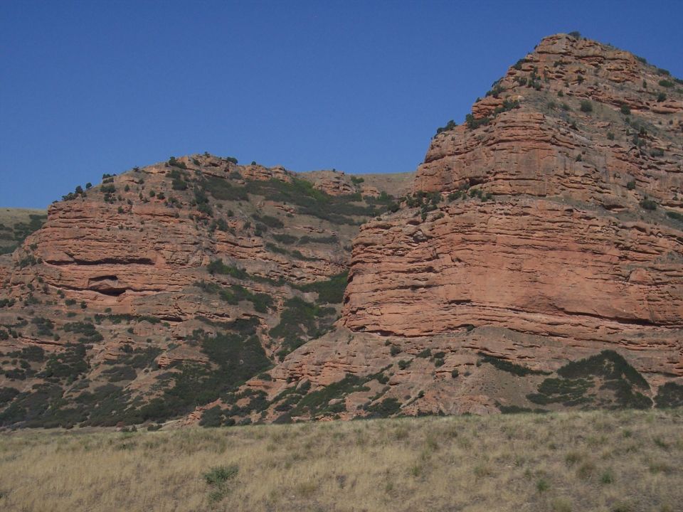 Rocky hills with layered red rock formations under a clear blue sky, with dry grass in the foreground