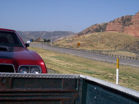 1972 Ford Ranchero - A red car with a black hood scoop is parked near a highway with a mountainous landscape in the background.