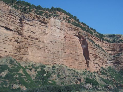1972 Ford Ranchero - A vast, layered red rock cliff dominates the landscape under a clear blue sky, with sparse green vegetation clinging to its slopes.