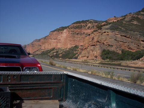 1972 Ford Ranchero - A red car travels down a highway with a scenic mountain backdrop under a clear blue sky.