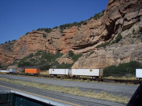 1972 Ford Ranchero - A train carrying shipping containers moves along tracks next to a large, cliff under a clear blue sky.