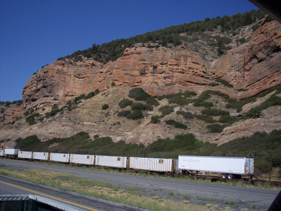 Train carrying multiple white shipping containers moves along tracks with a rocky, tree-covered mountain in the background