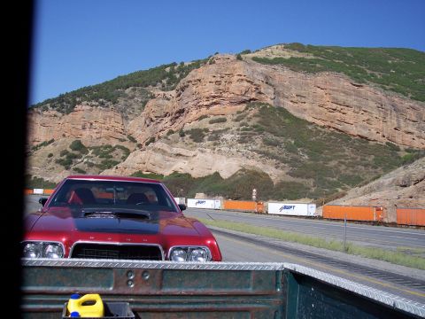 1972 Ford Ranchero - A red muscle car with a black hood scoop drives down a highway beside a long freight train.