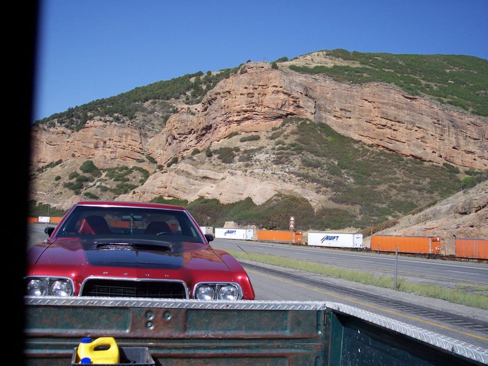 Red muscle car with black hood scoop driving on highway next to a train