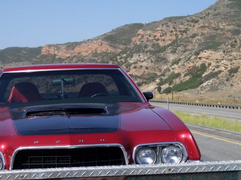 1972 Ford Ranchero - A red 1972 Ford Ranchero with a black hood scoop and diamond plate bumper is parked on the side of a highway with mountains in the backgrou…