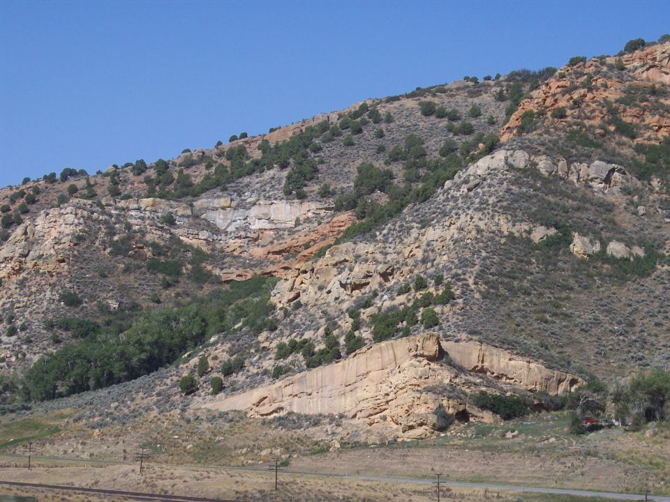 rocky hillside with scattered trees and bushes under a clear blue sky. A small red vehicle is visible on the right