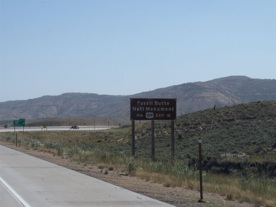 Roadside sign for Fossil Butte Natl Monument via 189 Exit 18, with distant mountains and a highway