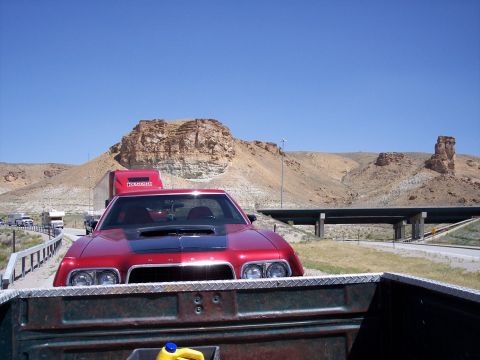 1972 Ford Ranchero - A red 1972 Ford Ranchero with a prominent black hood scoop is seen from the rear of a pickup truck, with a desert landscape and highway ove…