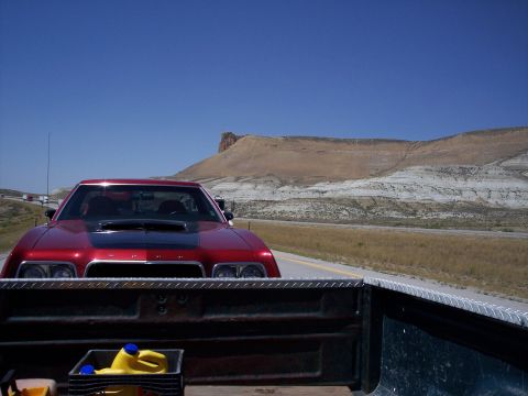 1972 Ford Ranchero - A red coupe with a black hood scoop is seen from the perspective of a truck bed on a highway with a large mountain in the distance.