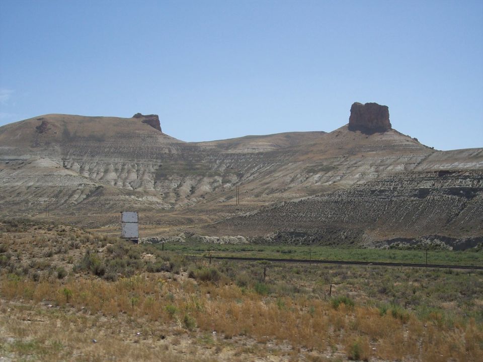Dry landscape with two mesas under a clear blue sky, a white billboard in the middle ground