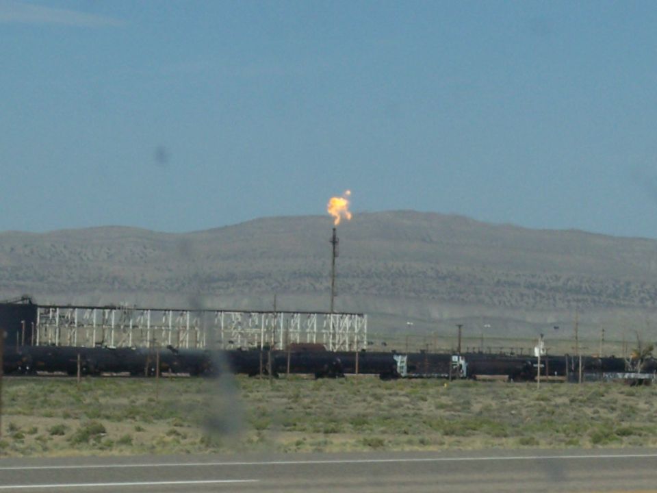 Train cars sit on tracks near an industrial facility with a flare stack burning