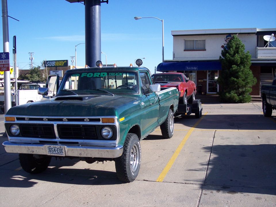 Green 1975 Ford F-Series pickup truck towing a red car on a trailer. Ford Truck decal on rear window