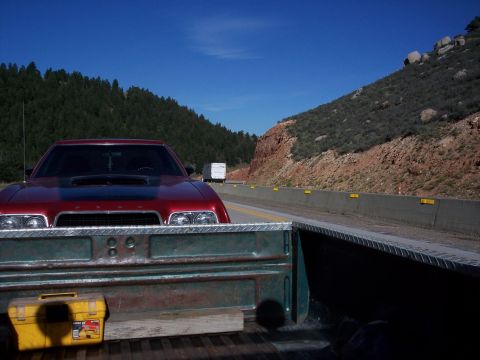 1972 Ford Ranchero - A red car with a black hood scoop is seen from the perspective of a truck bed on a highway.