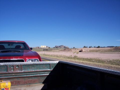 1972 Ford Ranchero - A red 1972 Ford Ranchero with a black hood scoop is seen driving on a highway, viewed from the bed of a pickup truck.