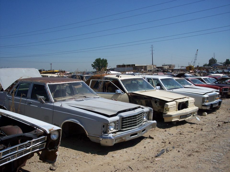 Several cars in a junkyard, front view. A light gray sedan with a rusty roof is in the foreground, its hood open