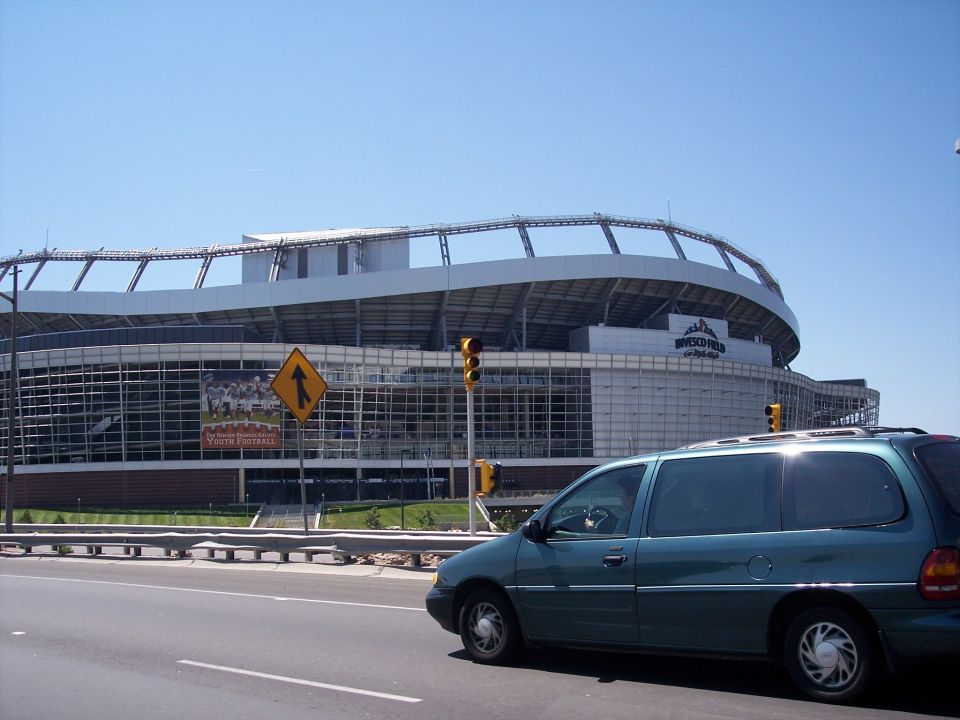 Teal minivan drives past a large stadium with 'BRONCOS FIELD' visible
