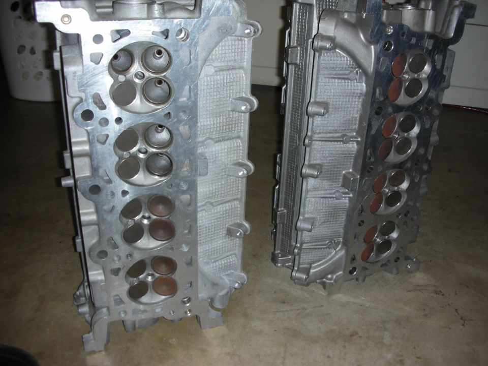 Two aluminum engine cylinder heads with valves visible, one on the left and one on the right