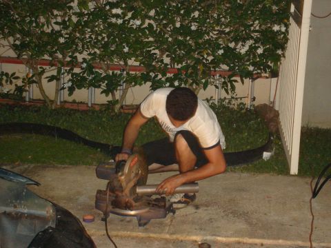 1995 Mazda MX-6 - Man cuts metal pipe with a chop saw outdoors, with a car fender visible on the left.