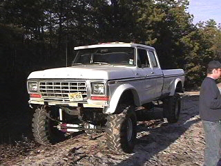 1987 Suzuki Samurai - White 1979 Ford F-150 pickup truck with large off-road tires and suspension lift parked on dirt.